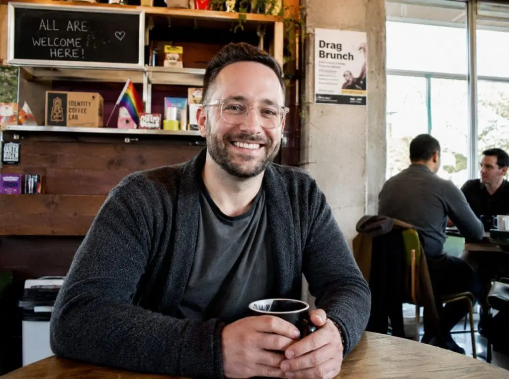 un homme de 30-45 ans souriant dans un café LGBTQ+ ou un lieu communautaire, ambiance chaleureuse et non cliché.
