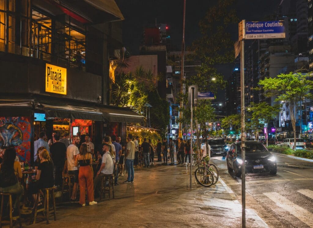 uma fotografia de uma rua movimentada de Pinheiros à noite, com bares, luzes urbanas e pessoas vistas de longe